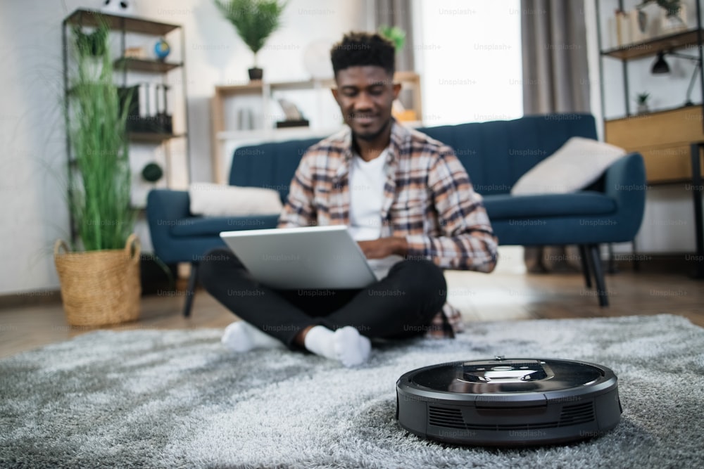 Positive african guy sitting on floor with laptop while robotic vacuum cleaner washing carpet at living room. Modern lifestyle with technology. Chores concept.