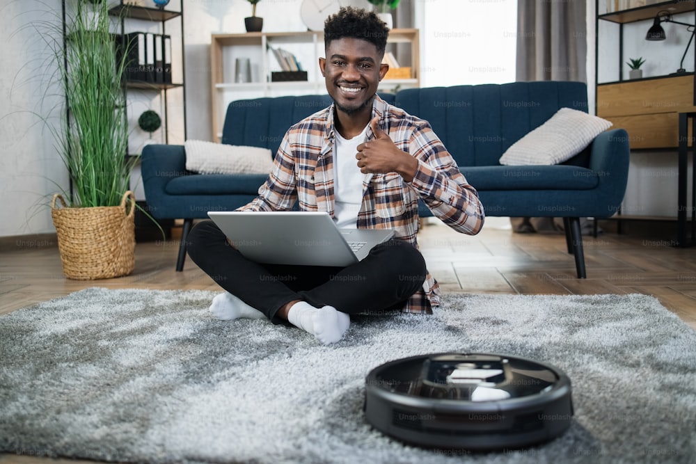 Young afro american guy showing thumb up for expressing his satisfaction with robot vacuum cleaner. Modern devices for chores.