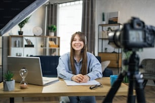 현대 카메라에 온라인 자습서를 녹화하는 헤드셋에서 행복한 백인 여성. 집에서 원격 근무를 하는 동안 노트북과 소프트 박스를 사용하는 웃는 여성.