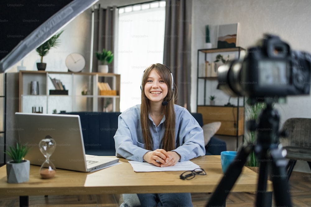 Donna caucasica felice in cuffia che registra tutorial online sulla fotocamera moderna. Femmina sorridente che utilizza laptop e soft box durante il lavoro a distanza a casa.