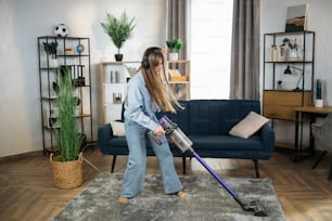 Charming young woman in modern headphones enjoying favorite music while cleaning house with hand held vacuum cleaner. Concept chores.