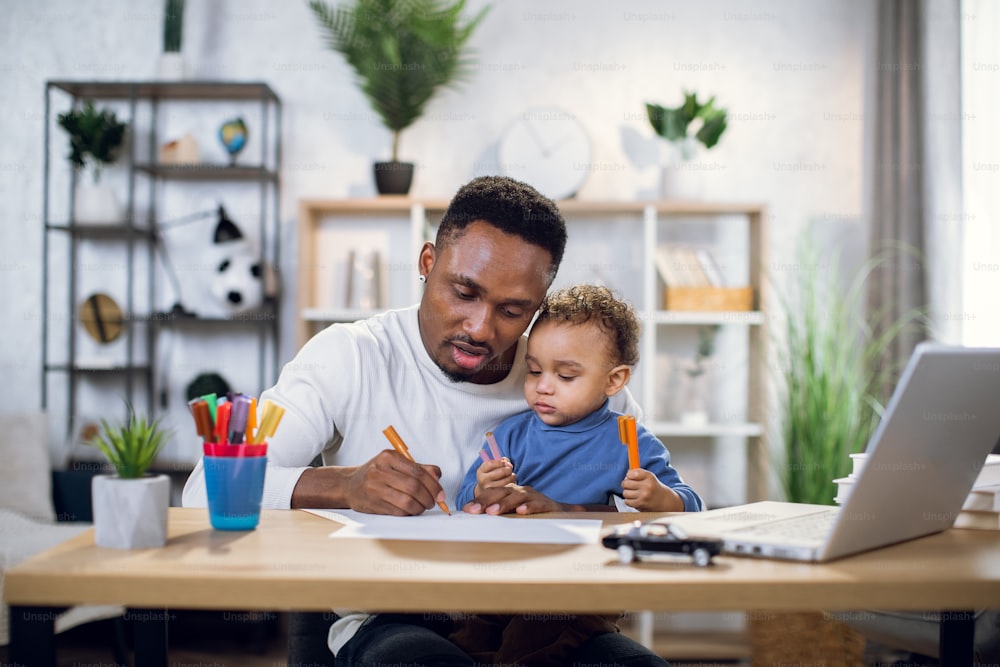 Handsome afro american man holding his cute son on knees and drawing with colorful pencils. Young father sitting at table with modern laptop and entertaining child.