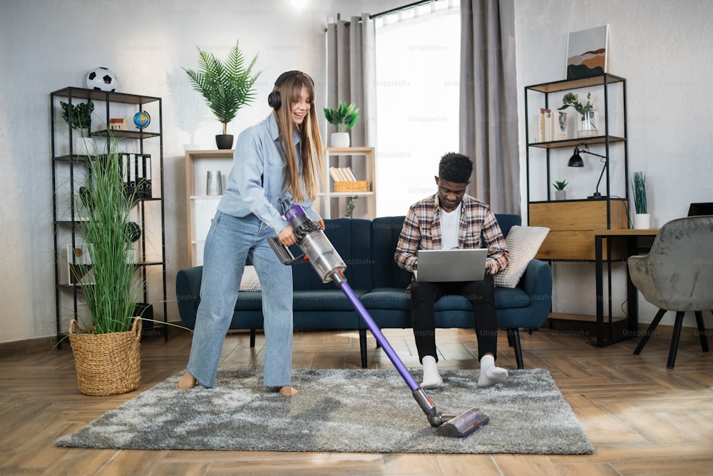 Happy caucasian woman listening music in wireless headphones while cleaning carpet with modern vacuum cleaner. Afro american man sitting on sofa and working on laptop.