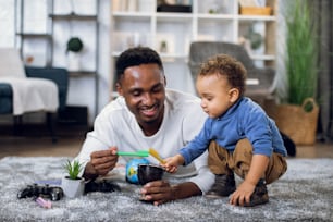 Young father and cute son drawing with pencils while lying together on carpet. Happy man and boy spending weekend time with creative activity.