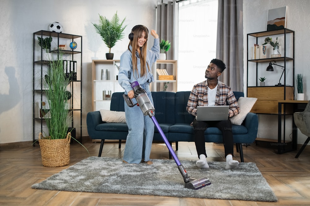Happy caucasian woman listening music in wireless headphones while cleaning carpet with modern vacuum cleaner. Afro american man sitting on sofa and working on laptop.