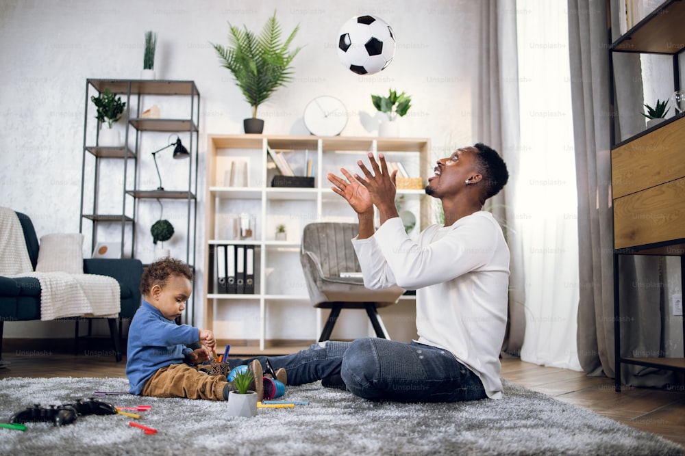 Felice padre nero in abbigliamento casual seduto con il suo figlioletto sul tappeto e giocando con la palla. Concetto di intrattenimento, tempo libero e famiglia.
