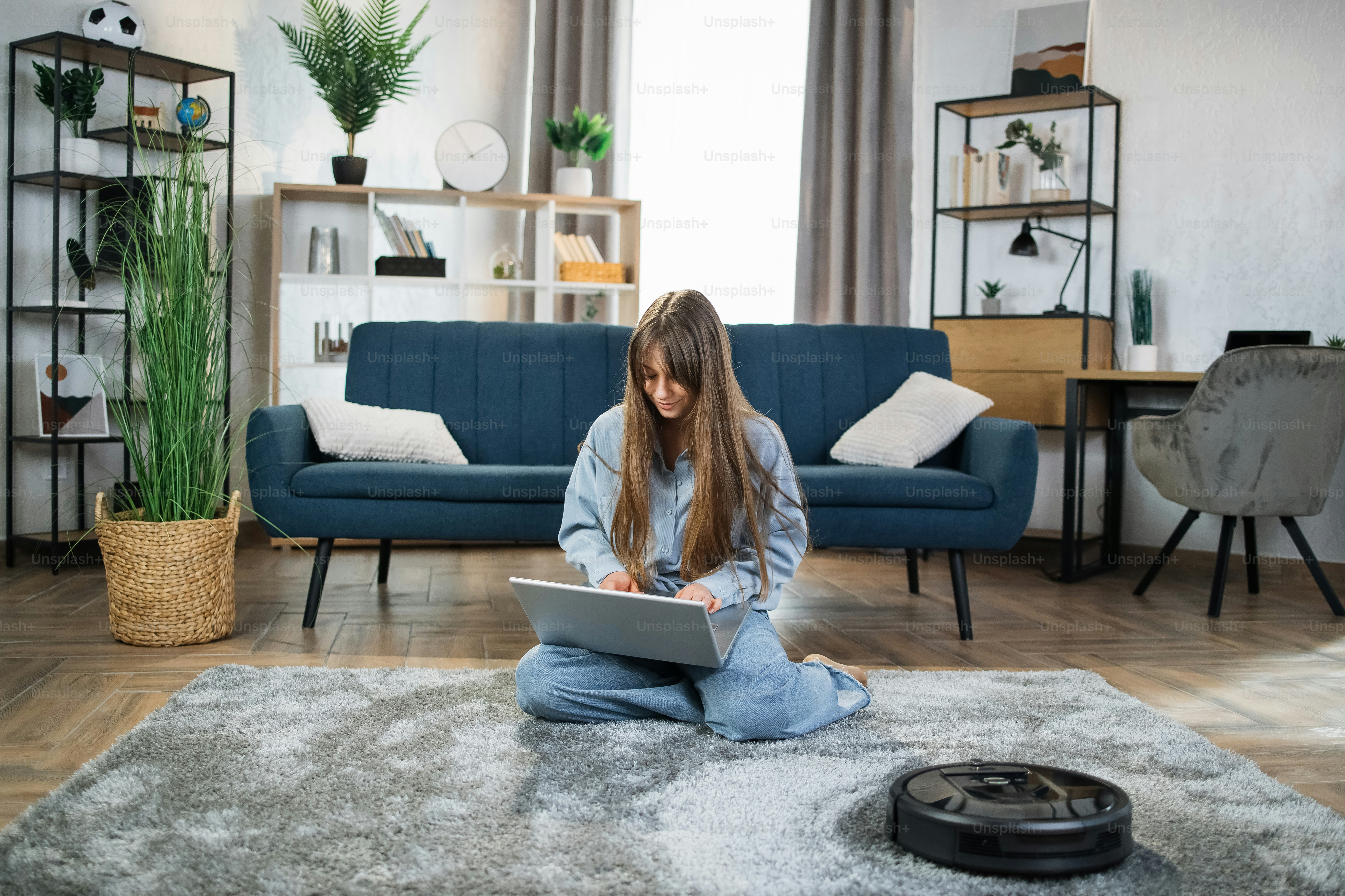 Schöne junge Frau, die auf dem Boden sitzt und am Laptop arbeitet, während der Staubsaugerroboter ihre Wohnung wäscht. Moderner Lifestyle mit Technologie