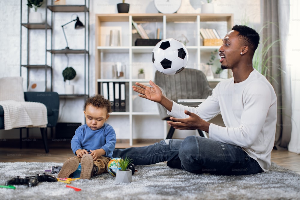 Glücklicher schwarzer Vater in Freizeitkleidung, der mit seinem kleinen Sohn auf dem Teppich sitzt und mit dem Ball spielt. Konzept von Unterhaltung, Freizeit und Familie.