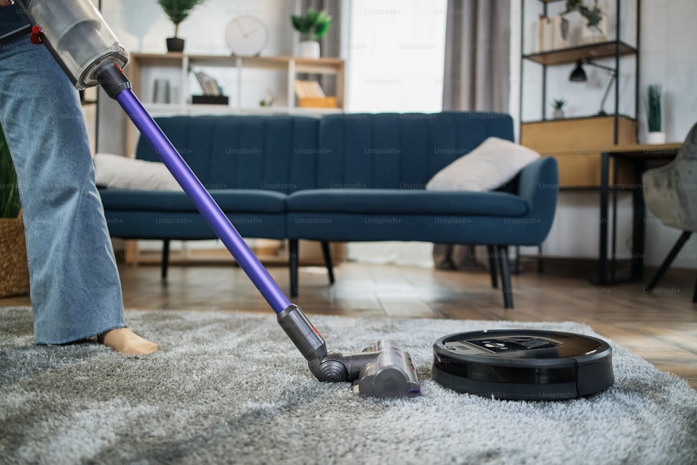 Close up of young woman cleaning carpet with hands vacuum cleaner while another wireless robot washing floor. Concept of technology and households.