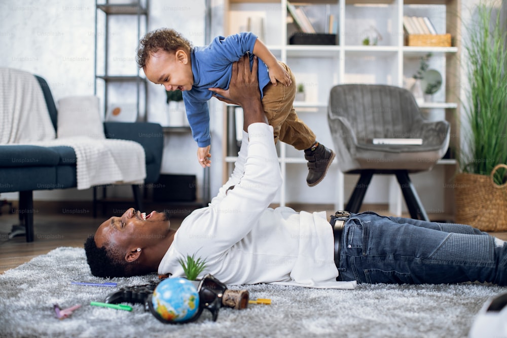 Fröhlicher afrikanischer Mann, der auf einem weichen Teppich liegt und mit einem kleinen Jungen spielt. Junger Vater und Sohn genießen die gemeinsame Zeit zu Hause. Familienkonzept.