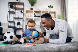 Afro american father and cute little son having fun with his cute son during leisure time at home. Young man and baby boy playing with various toys on carpet.