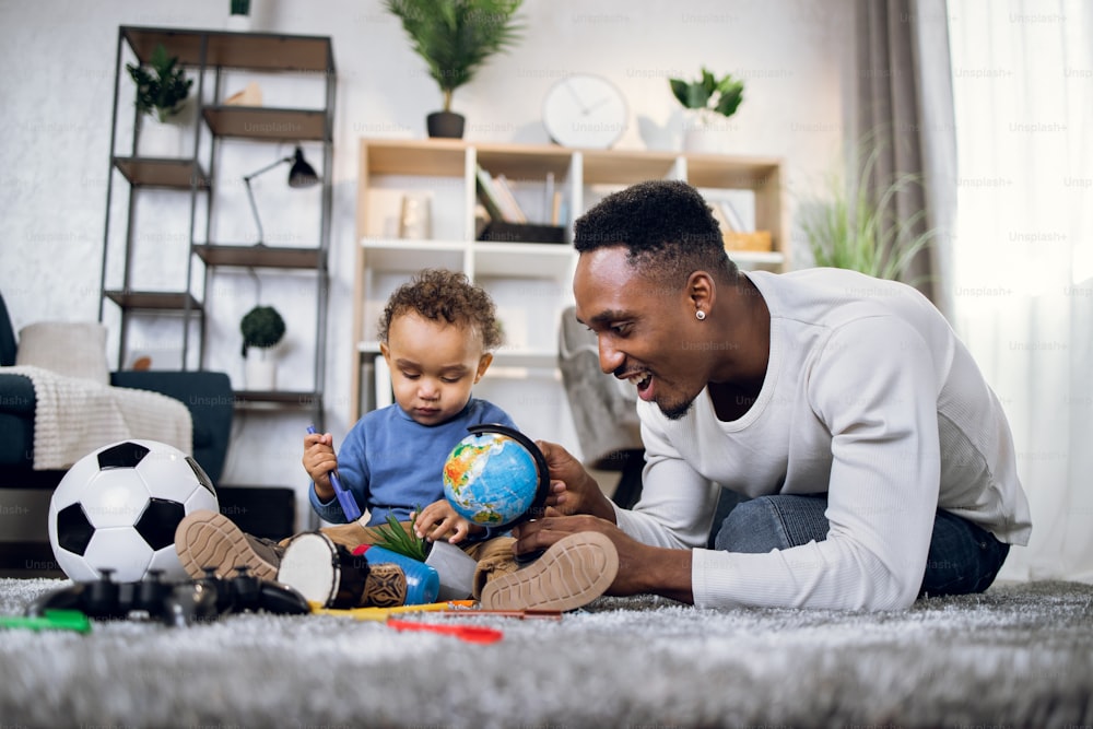Afroamerikanischer Vater und süßer kleiner Sohn, der Spaß mit seinem süßen Sohn in der Freizeit zu Hause hat. Junger Mann und kleiner Junge spielen mit verschiedenen Spielsachen auf Teppich.