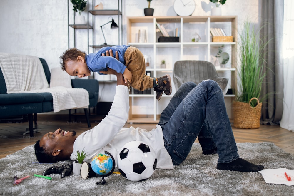 Homem africano alegre deitado no tapete macio e brincando com o menino. Pai e filho jovens desfrutando do tempo passando juntos em casa. Conceito de família.