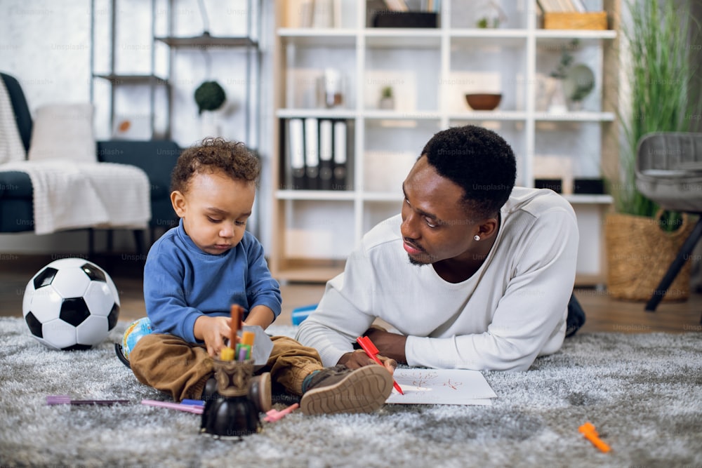 Padre afroamericano amoroso e hijo lindo acostado juntos en el piso y dibujando con lápices de colores en el álbum. Tiempo en familia en casa. Concepto de unión.