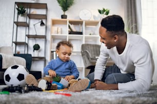 Young afro american father taking care of his cute son while staying together at home. Happy man and baby boy in casual clothes sitting on carpet and playing with toys.