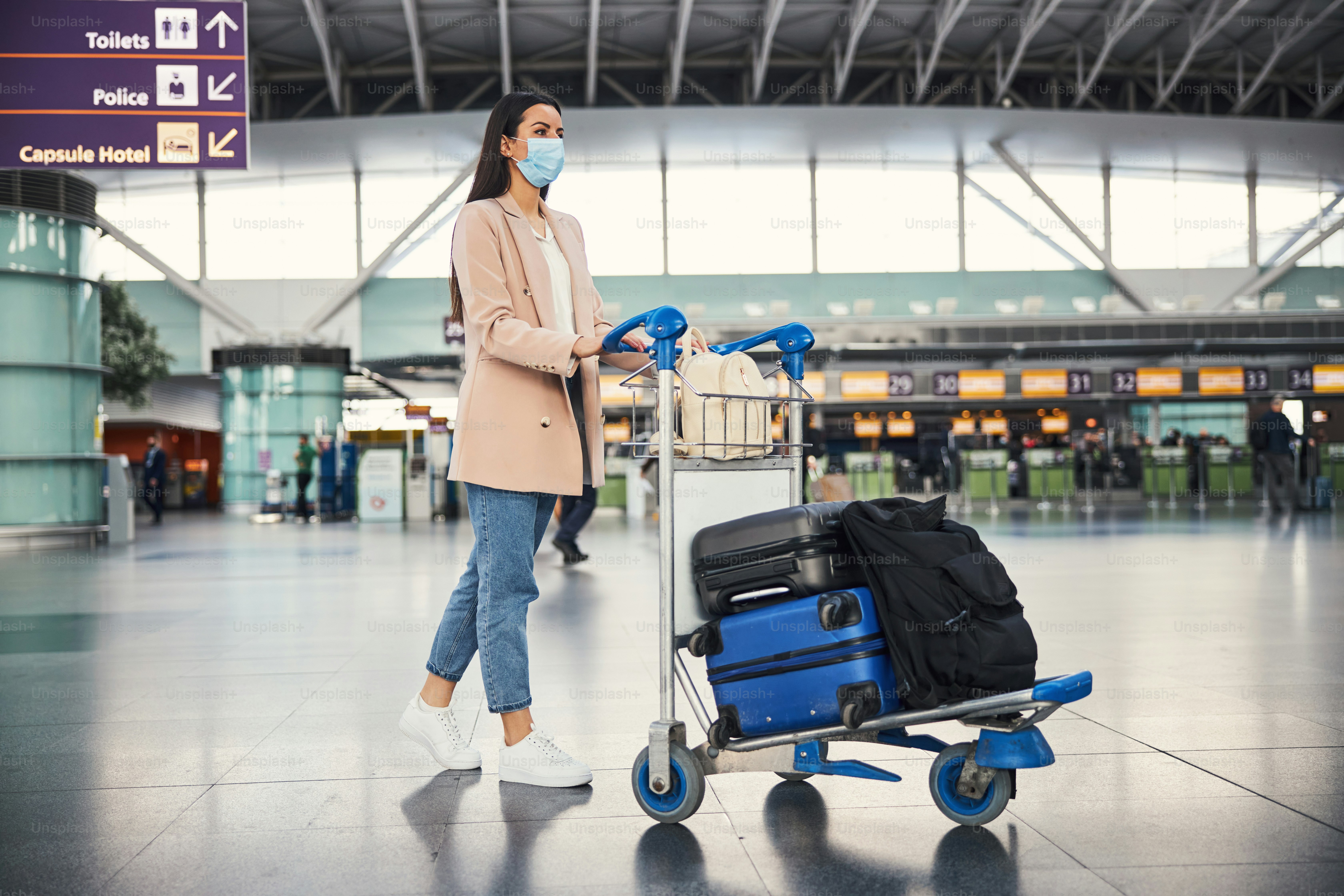 Elegant lady wearing protective face mask while walking down airport terminal with luggage trolley