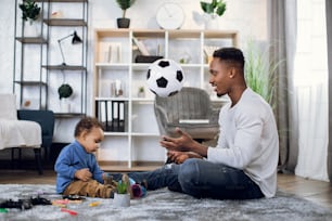 Happy black father in casual wear sitting with his little son on carpet and playing with ball. Concept of entertainment, leisure time and family.