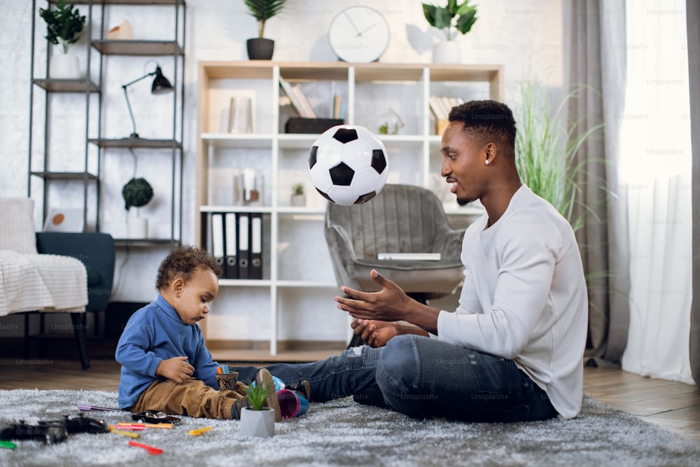 カジュアルな服を着た幸せな黒人の父親がカーペットの上に小さな息子と一緒に座り、ボールで遊んでいます。娯楽、余暇、家族のコンセプト。