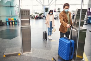 Pasajero masculino con mascarilla médica comprobando el peso del equipaje mientras una mujer hace cola a una distancia segura