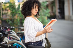 Black adult woman reading story book in downtown city. Woman standing on street and reading book.