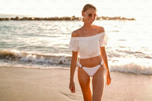 Hermosa mujer caucásica sonriente en un paseo relajante en la playa vacía en la orilla del mar al atardecer. Vibraciones veraniegas.