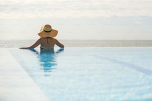 Beautiful woman in hat relaxing at edge in blue pool, enjoying summer vacation and sea view. Slim young female relaxing at tropical resort in swimming pool. Travel and Holidays. Space for text