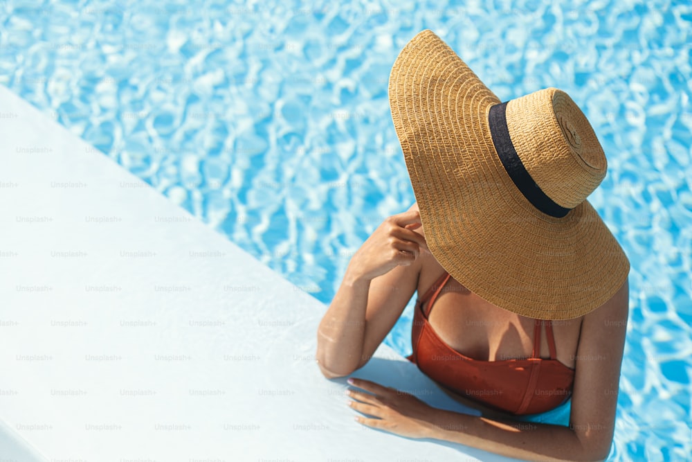 Beautiful woman in hat relaxing in pool water, enjoying summer vacation at tropical resort. Slim young female sunbathing at swimming pool edge, view above. Holidays and travel. Space for text