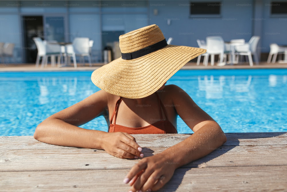 열대 리조트에서 여름 휴가를 즐기고 있습니다. 수영장 나무 부두에서 물속에서 휴식을 취하는 모자를 쓴 아름다운 여자. 수영장 가장자리에서 일광욕을 하는 날씬한 젊은 여성, 위의 전망. 휴일 및 여행