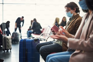 Stylish man in medical mask checking boarding pass while woman using hand sanitizer in departure lounge
