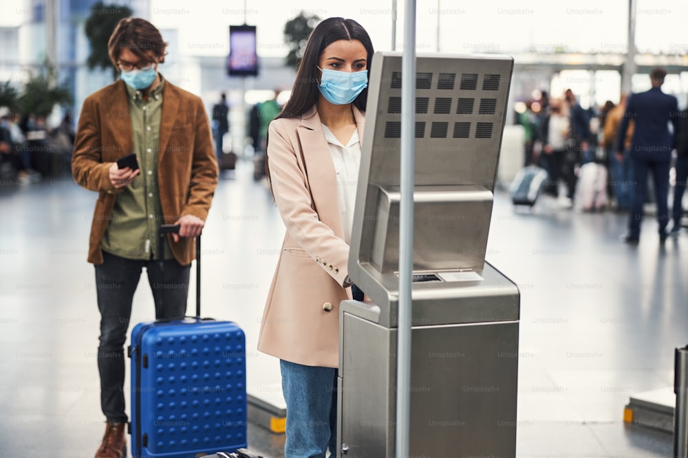 Elegant female passenger in medical mask checking luggage weight while man standing in line and using smartphone