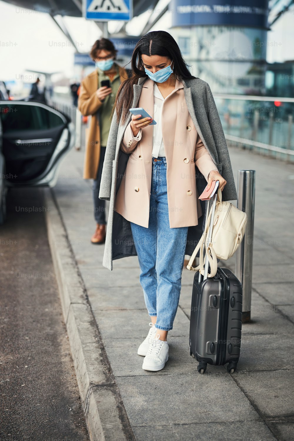 Signora elegante con valigia da viaggio che indossa una maschera protettiva per il viso mentre legge il messaggio sullo smartphone per strada