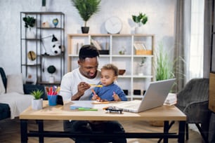 Afro american man working on modern laptop and playing with baby boy while sitting at table. Multitasking father trying to work remotely from home.