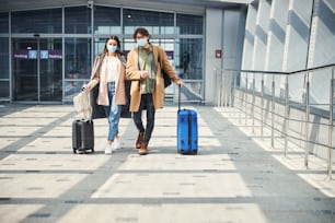 Stylish man and woman with travel suitcases wearing protective face masks while waiting for the flight at airport