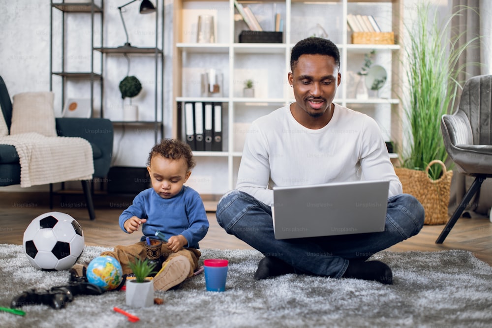 Afro american baby boy sitting on carpet and playing with toys while his father sitting near and working on laptop. Young man carrying baby and doing remote job.