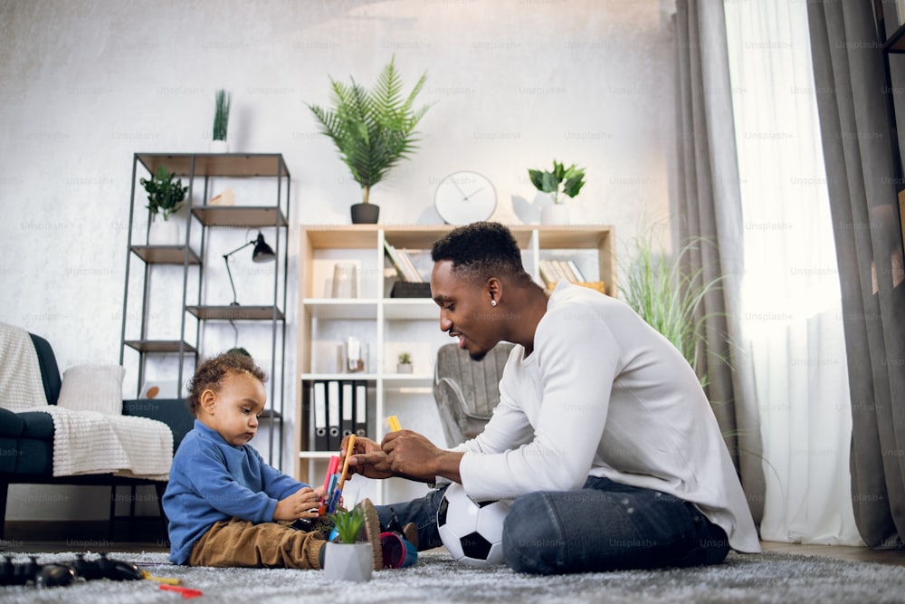 Side view of afro american father and cute little son using various toys while playing together on soft carpet. Concept of parenting and leisure time.