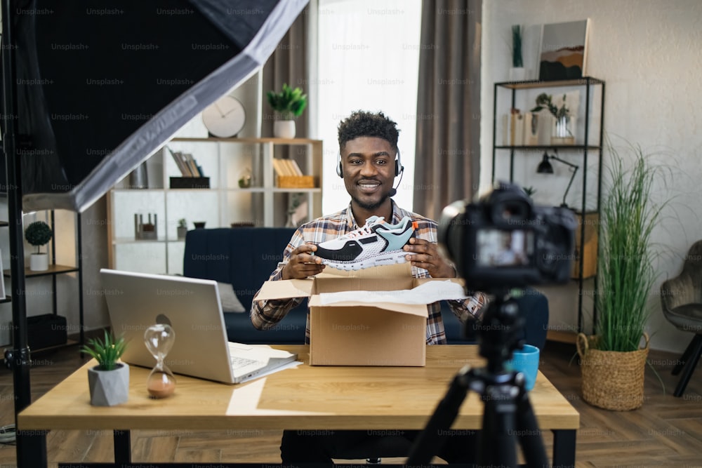 Uomo africano alla moda che fa il disimballaggio di nuove scarpe da ginnastica durante la registrazione di video sulla telecamera. Concetto di persone, blogging e pubblicità.