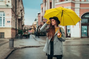 Joven hermosa mujer esperando un taxi y sosteniendo un paraguas amarillo mientras está en la ciudad en un día lluvioso