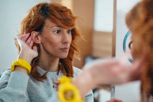 Junge rothaarige Frau steckt sich ein Hörgerät ins Ohr, während sie sich selbst im Spiegel betrachtet