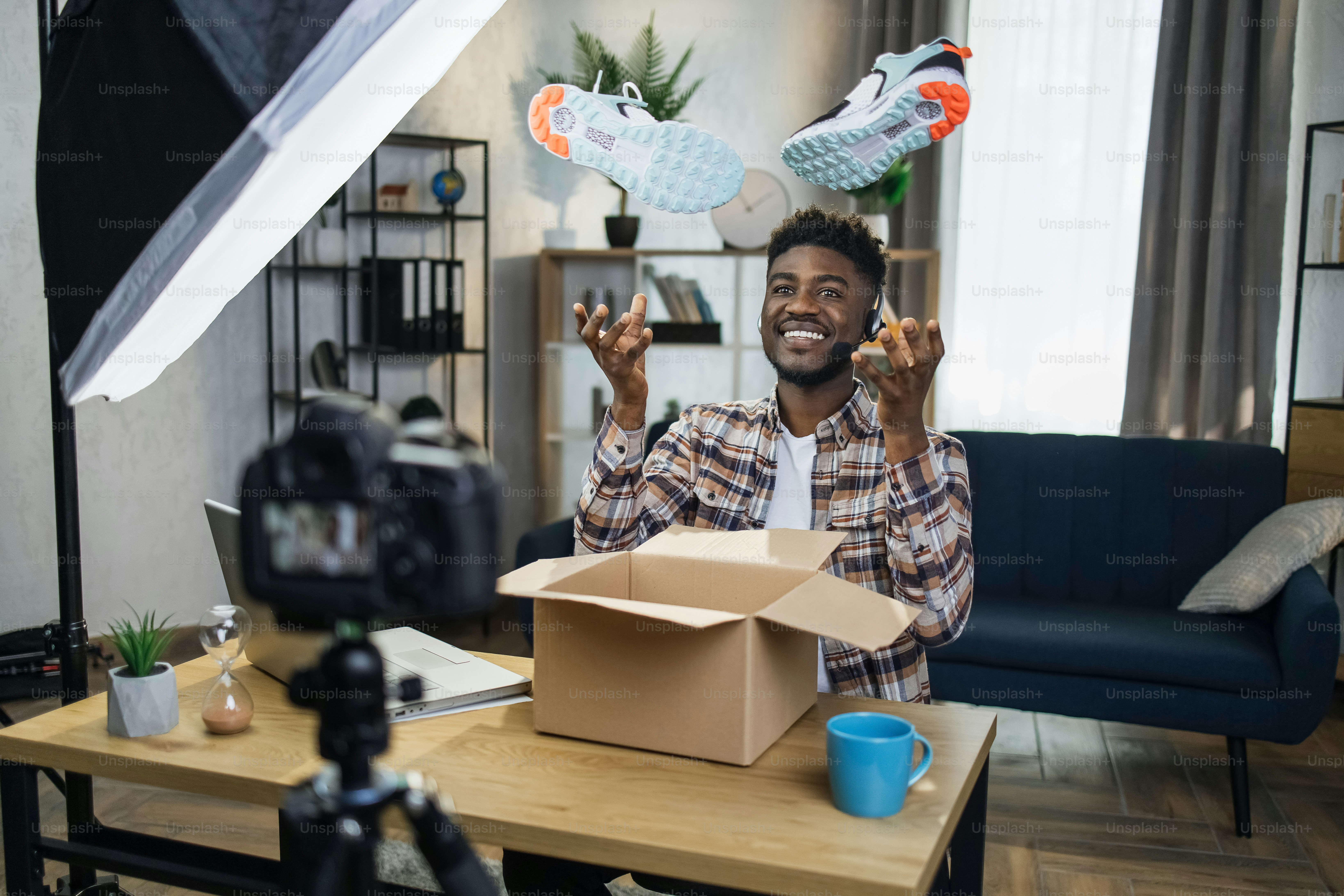 Alegre hombre afro con auriculares creando contenido de video para sus redes sociales mientras desempaca zapatillas nuevas en casa. Bloguero feliz que disfruta de su pasatiempo favorito con la ayuda de dispositivos modernos.