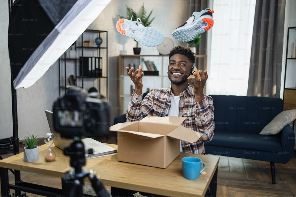 Homme afro joyeux dans un casque créant du contenu vidéo pour ses réseaux sociaux tout en déballant de nouvelles baskets à la maison. Blogueur heureux profitant de son passe-temps préféré à l’aide de gadgets modernes.