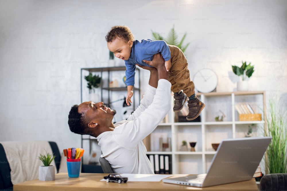 Cheerful african man playing with cute baby boy and using modern laptop for remote work. Young father working from home and taking care of son.