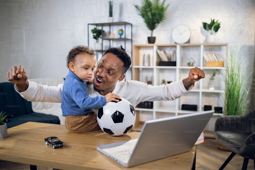 Joyful african man and little boy sitting together at table and using modern laptop for watching football match. Father and son spending time at home.