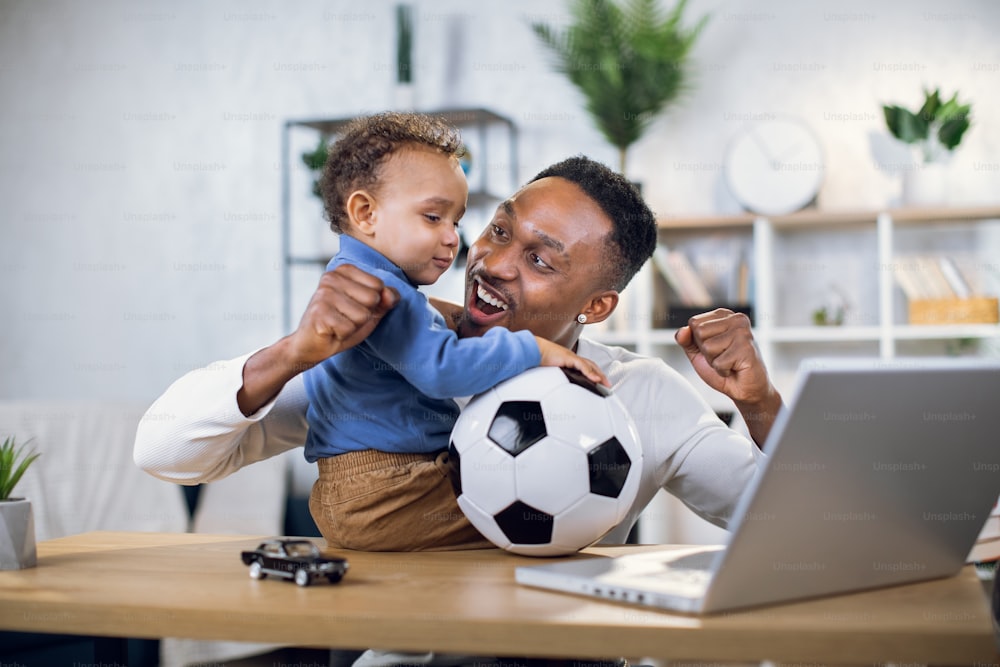 Cheerful african father and his cute son using modern laptop for watching football game at home. Concept of parenting, technology and leisure time.