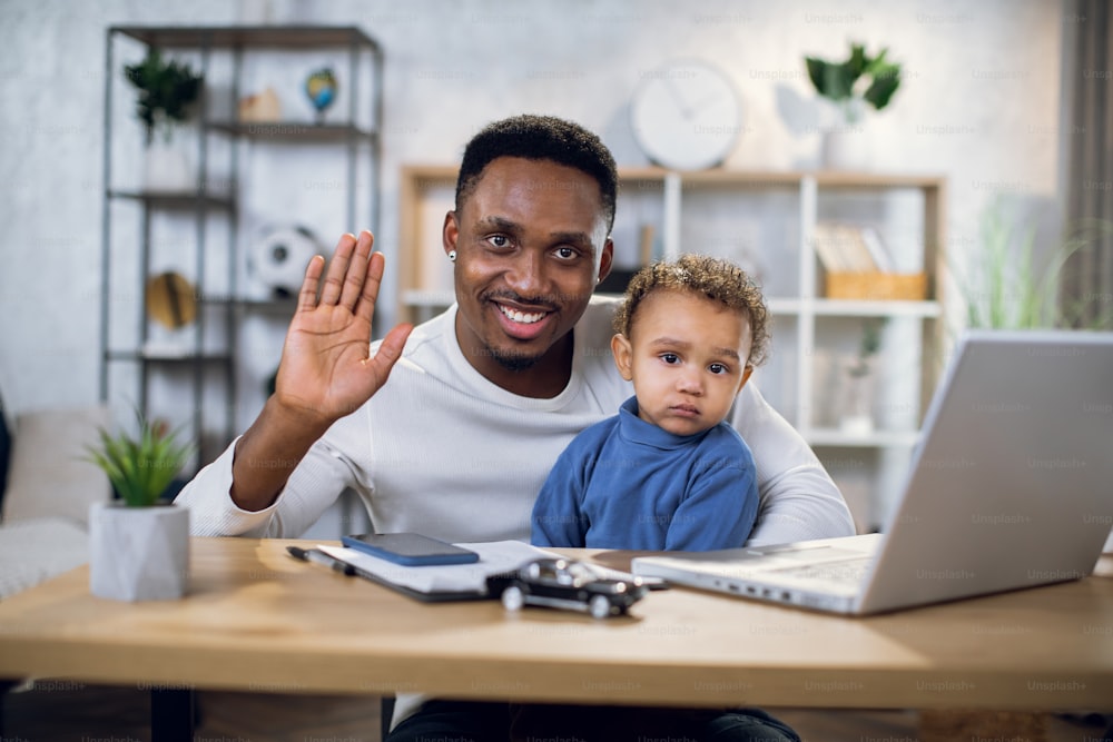 Smiling african man waving on camera while sitting at home office with his cute baby boy on knees. Concept of parenting, technology and freelance.