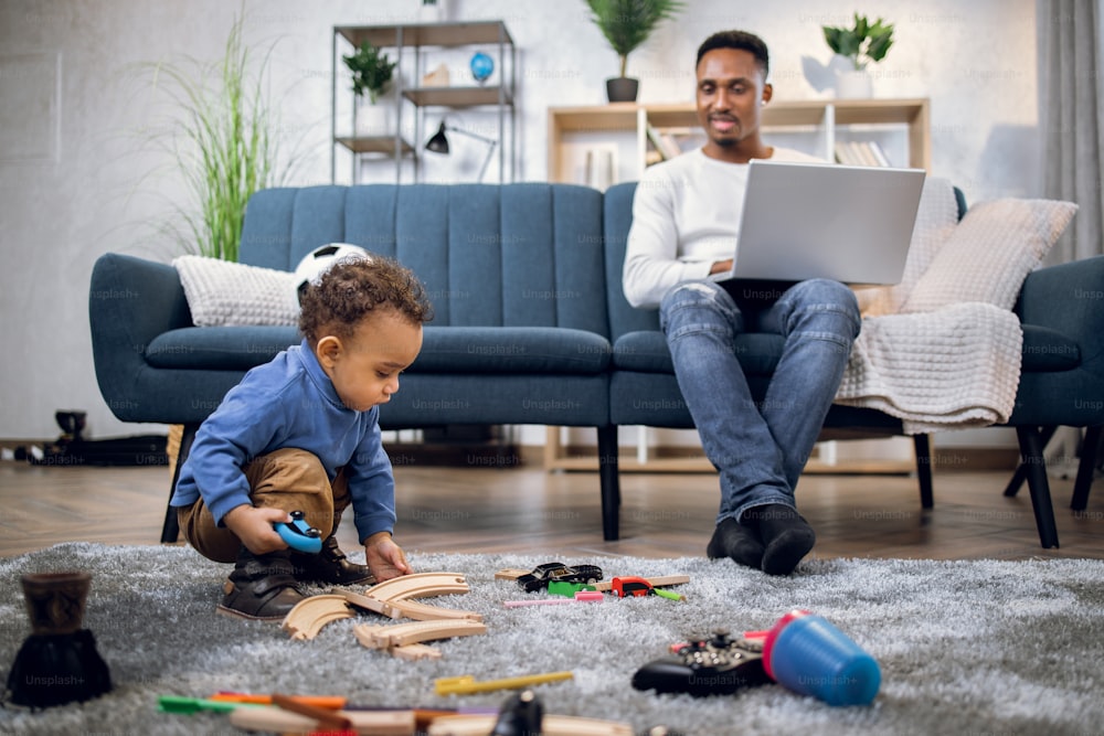 African young man sitting on couch and working on wireless laptop while little baby boy playing with toys on carpet. Concept of parenting and freelance.