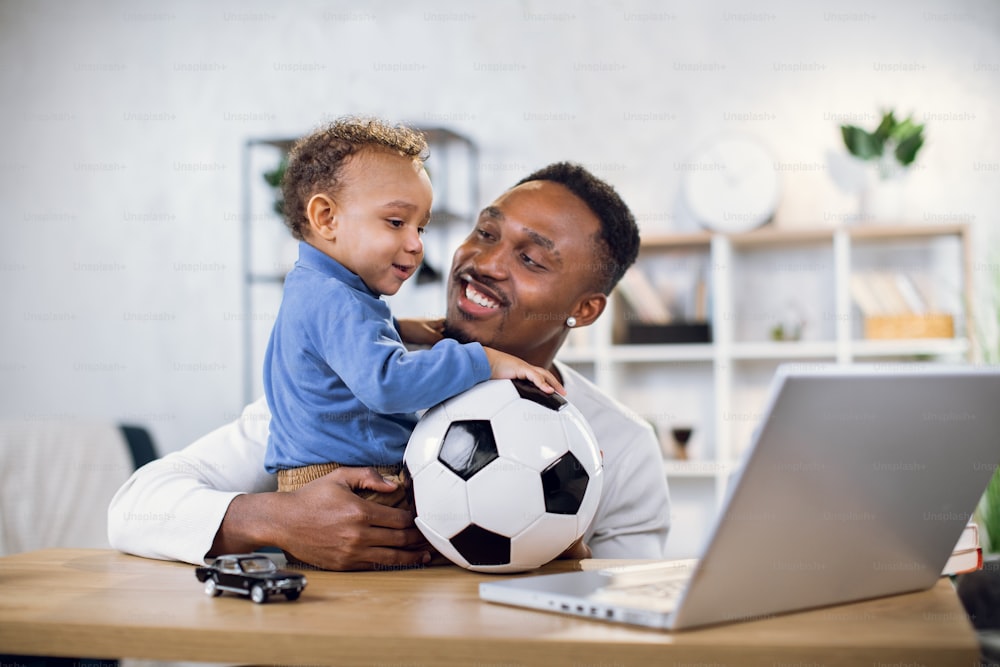 Young father and cute son playing with ball and watching something on modern laptop. Afro american man and little boy sitting at table and having fun together.