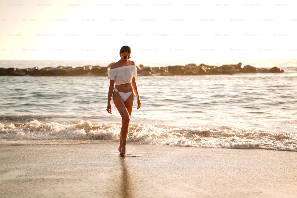 Hermosa mujer caucásica en un relajante paseo por la playa vacía en la orilla del mar al atardecer. Vibraciones veraniegas.