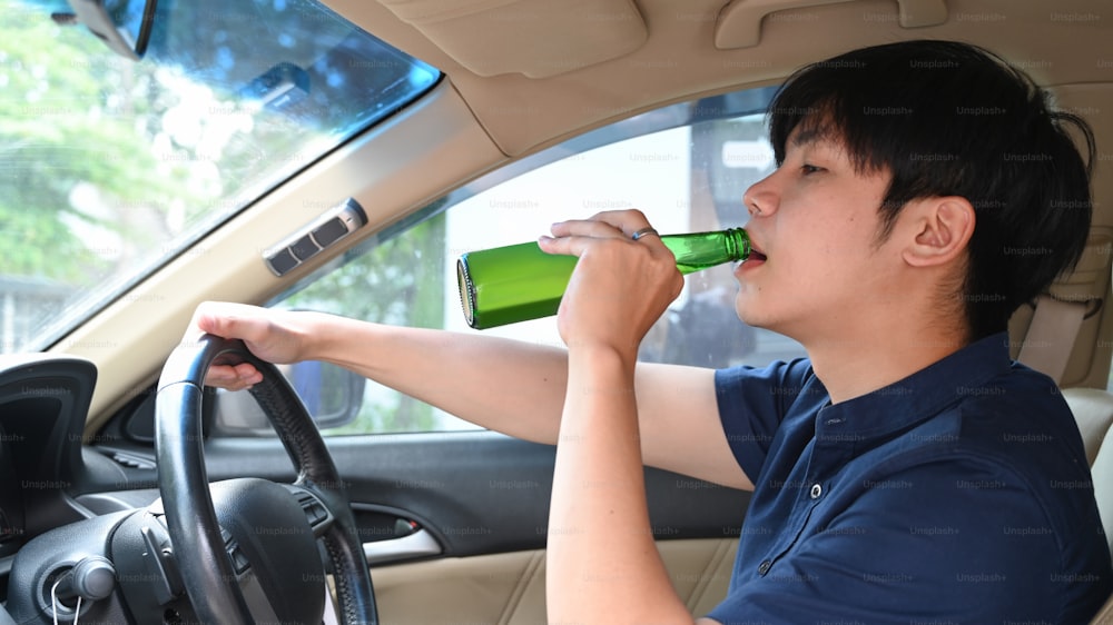 Young asian man drinking beer while driving a car. Driving under the influence.