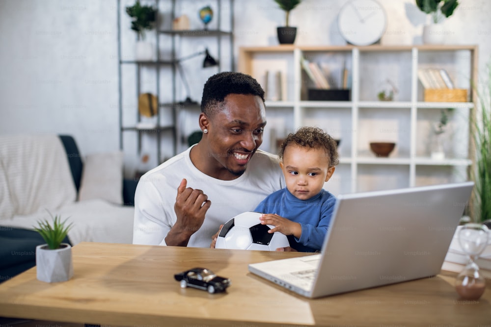 Uomo afroamericano e ragazzino che guardano la partita di calcio sul laptop wireless mentre si siedono insieme al tavolo. Padre e figlio felici che trascorrono il tempo libero utilizzando la tecnologia moderna.
