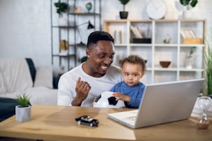 Afroamerikanischer Mann und kleiner Junge, der Fußballspiel auf einem drahtlosen Laptop schaut, während er zusammen am Tisch sitzt. Glücklicher Vater und Sohn verbringen Freizeit mit moderner Technologie.
